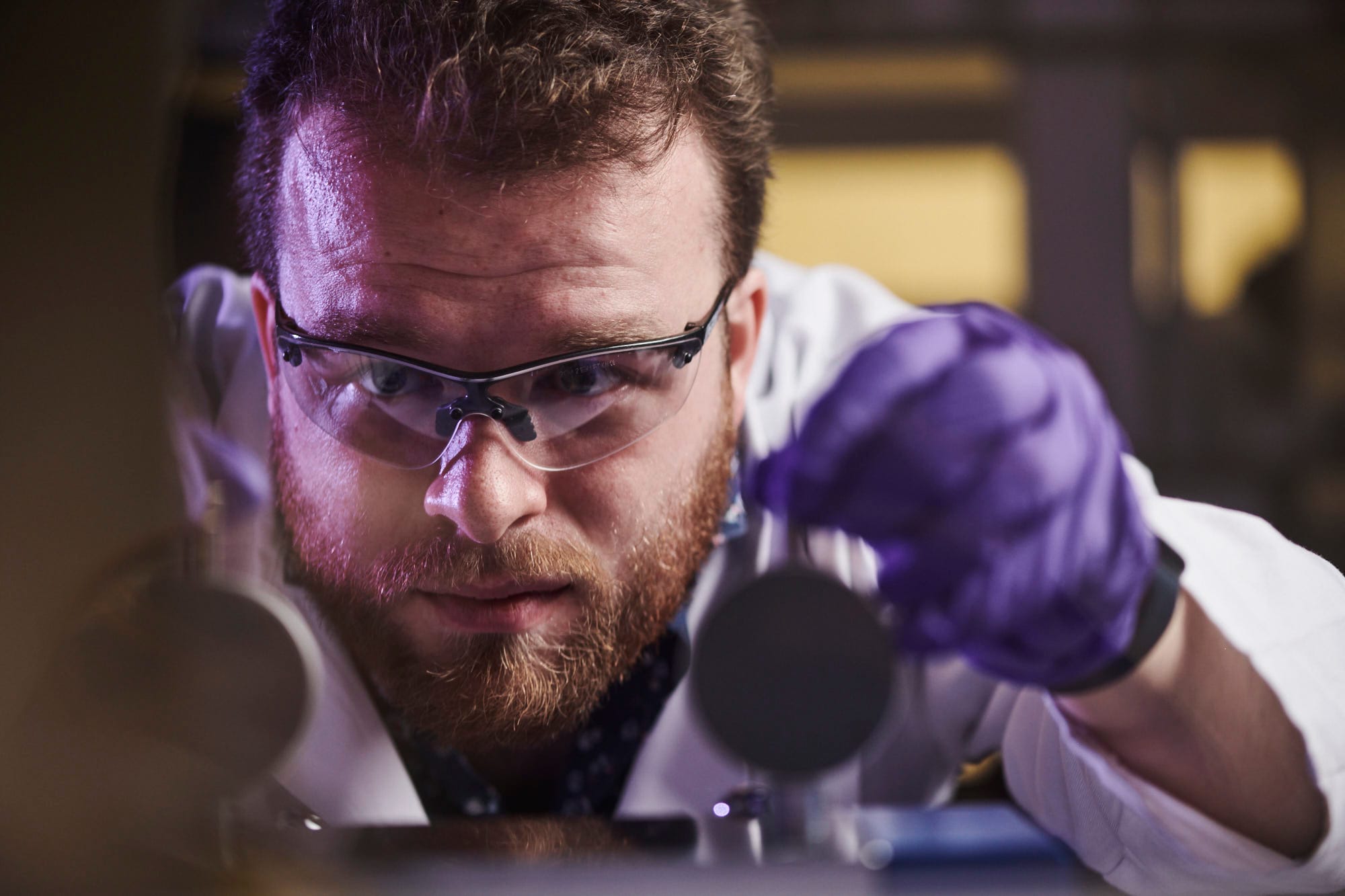 Close-up of a male scientist wearing safety glasses and purple gloves, carefully handling battery components in a lab, focused on lithium-ion battery research for electric vehicles