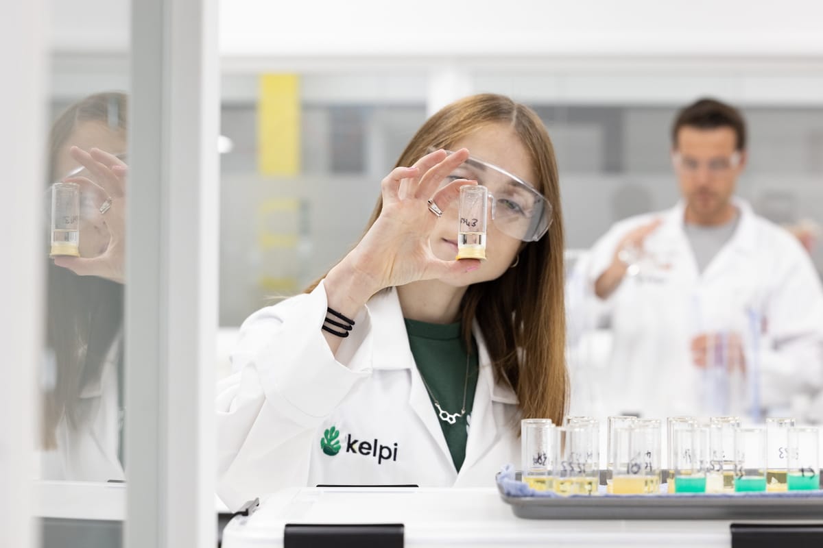 A scientist wearing safety goggles and a white lab coat with a Kelpi logo examines a vial of liquid in a laboratory setting