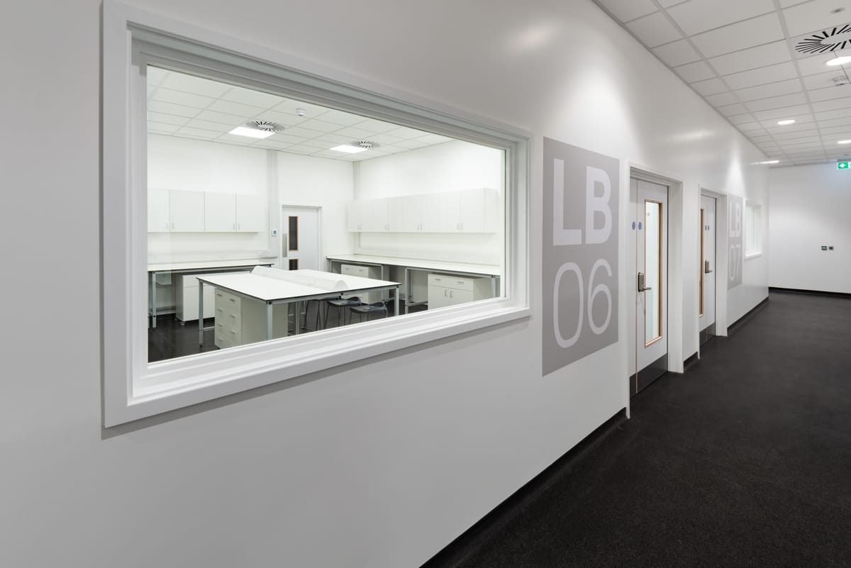 View through a large internal window into a clean, white laboratory room labeled LB06 at the Science Creates Old Market incubator, featuring lab benches, storage cabinets, and stools.