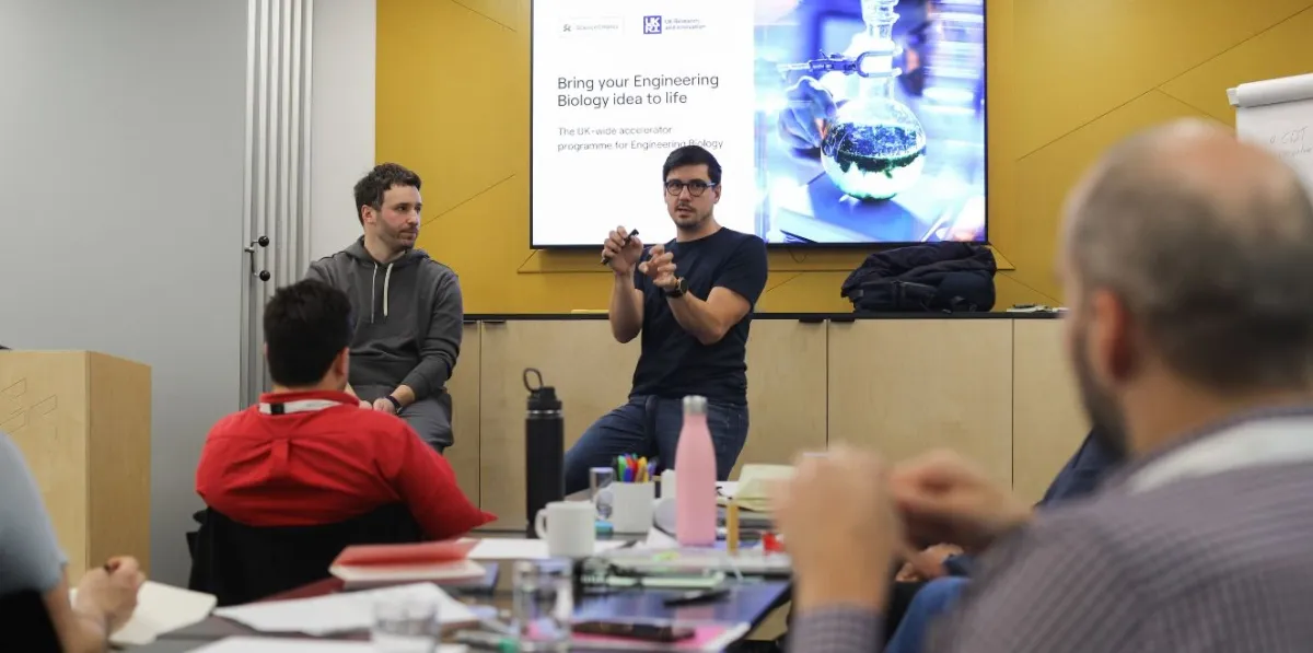 A group of people sit around a table during a talk about the Science Creates Engineering Biology Accelerator.