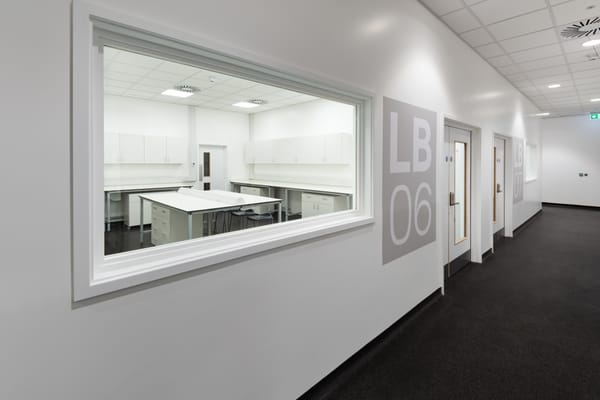 View through a large internal window into a clean, white laboratory room labeled LB06 at the Science Creates Old Market incubator, featuring lab benches, storage cabinets, and stools.