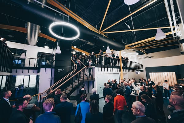 A large group of people gathered inside the Science Creates Old Market incubator during its 2022 launch event. Attendees stand on both floors, listening to a speaker on the staircase.